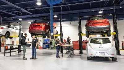 Wide view of a very modern automotive garage with cars on lifts. Instructor speaks to students.