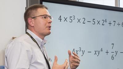 Instructor speaking next to a large digital screen with mathematic equations.