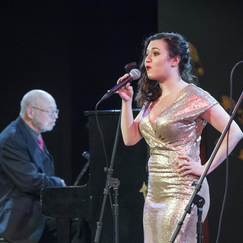 Woman on stage sings into a microphone with pianist in the background.