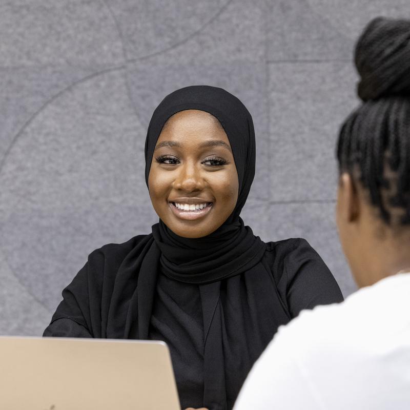 Woman in headscarf at a laptop computer smiles at client seated across from her.