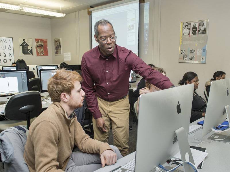 An instructor works with a student at a computer in a classroom.