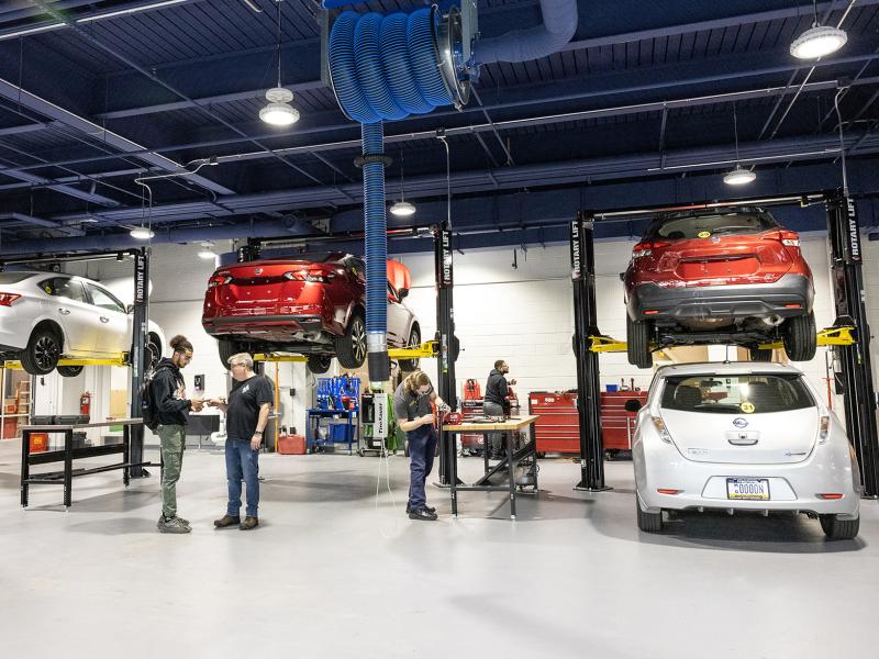 Wide view of a very modern automotive garage with cars on lifts. Instructor speaks to students.