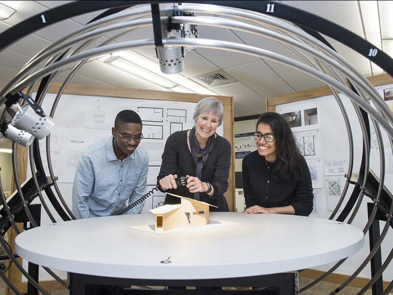Two students working with a smiling professor as they light an architectural model with professional equipment