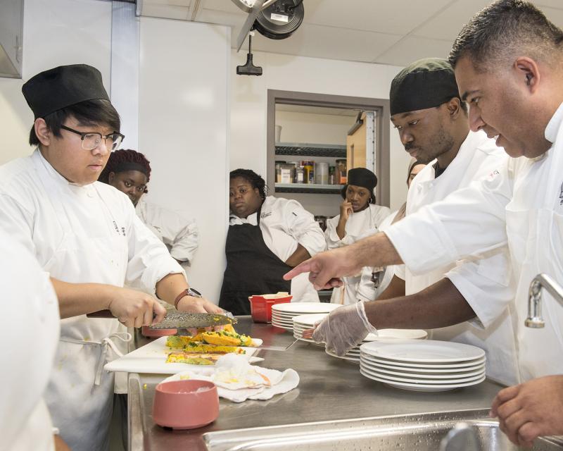 Culinary arts student slicing food as their teacher and other students watch