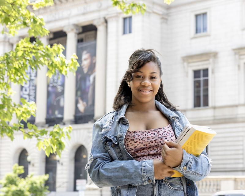 A student smiles and stands outside the mint building on a sunny day.