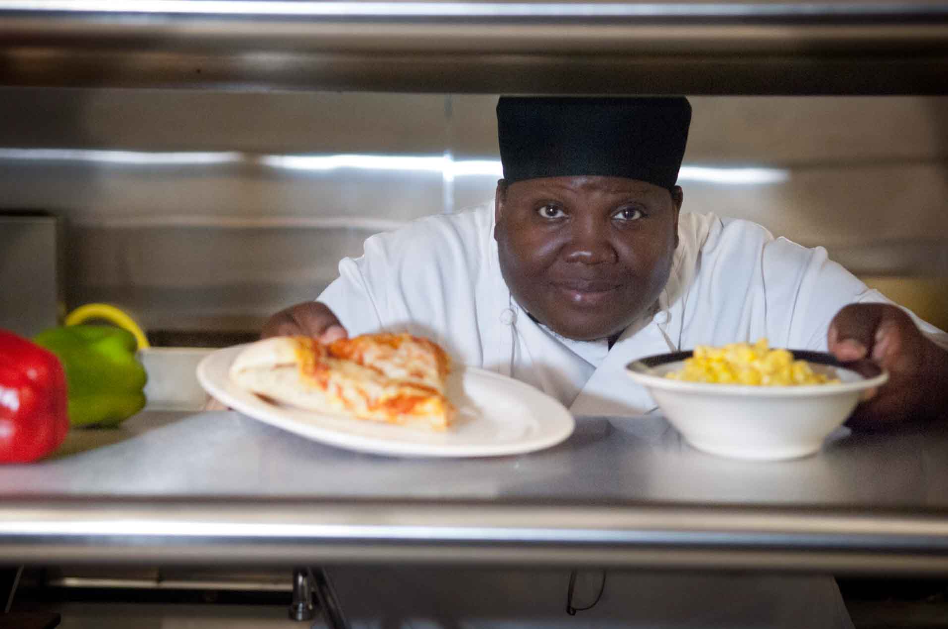 A woman in chef's clothing placing pizza and corn on a counter.