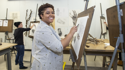 smiling student in art studio classroom drawing with charcoal at an easel