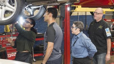 Instructor and two students watch another student working under a car on a lift.
