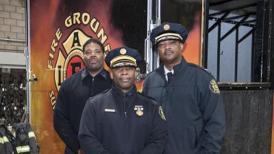Three fire figthers in dress uniforms pose for the camera with equipment in the background.