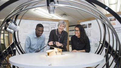 Two students working with a smiling professor as they light an architectural model with professional equipment