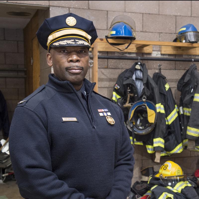 Fireman poses for the camera with helmets and coats hanging in the background.