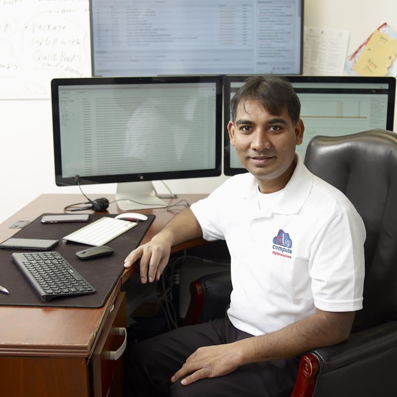 A network administrator sitting at a desk with three computer screens smiles for the camera.