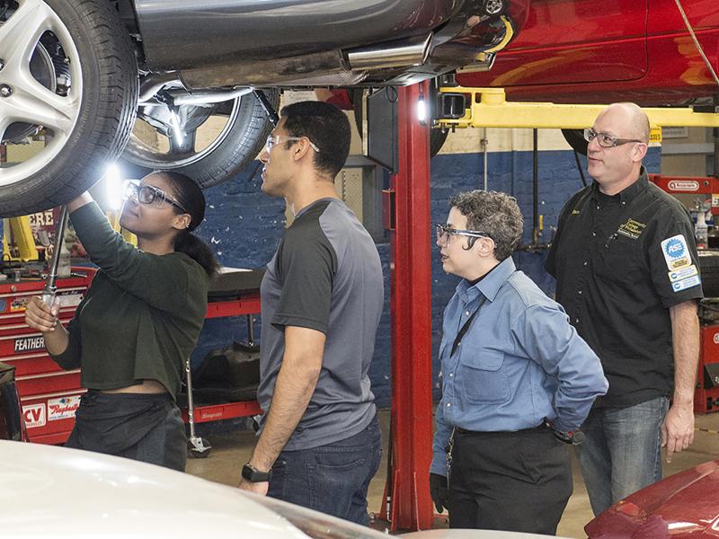 Instructor and two students watch another student working under a car on a lift.