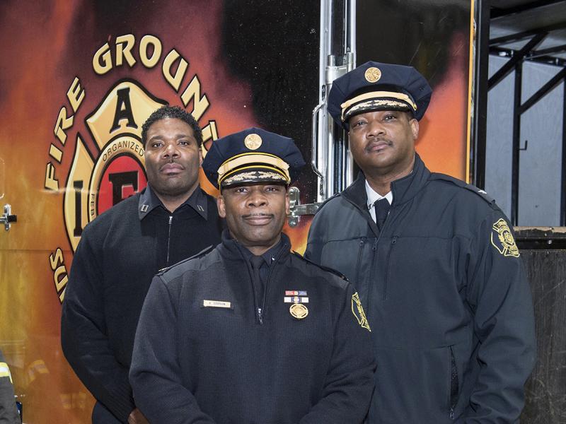 Three fire figthers in dress uniforms pose for the camera with equipment in the background.