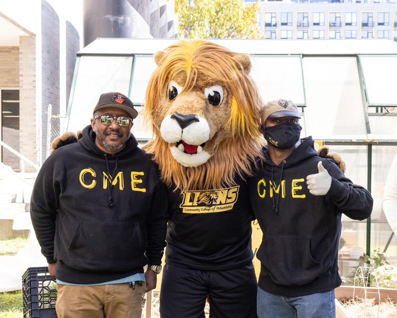 Roary in a greenhouse with two Students wearing Center for Male Engagement Hoodies