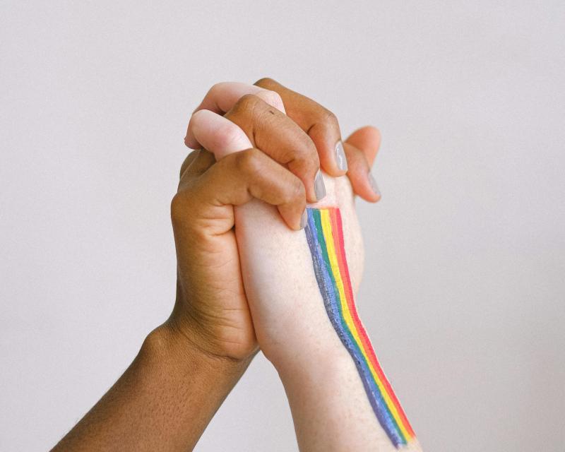 two diverse women's hands interlocked with gay flag painted on one hand
